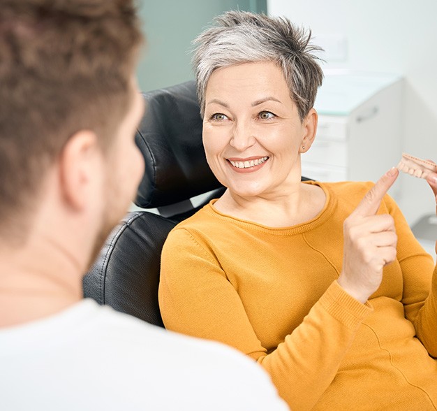 A smiling older woman sitting in a dentist’s chair while pointing to a denture she’s holding